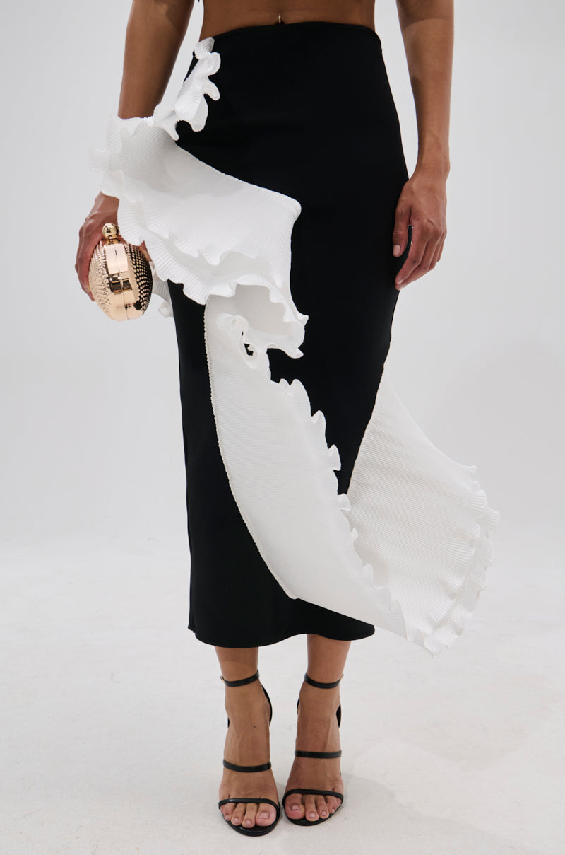 A person wearing the FASHION ICON RUFFLE BANDAGE MAXI SKIRT featuring bold white ruffle details, paired with a round metallic clutch bag and black strappy high-heeled sandals. 2