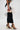 A person showcases the FASHION ICON RUFFLE BANDAGE MAXI SKIRT—a black pleated ruffle skirt with white ruffle accents—paired with black and gold heeled sandals and a small gold mesh clutch, set against a plain white background.