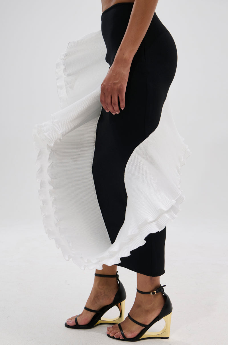 A person wears the FASHION ICON RUFFLE BANDAGE MAXI SKIRT, featuring a dramatic white pleated ruffle detail over black leggings, paired with black open-toe heels with gold block heels. The background is plain white. 4