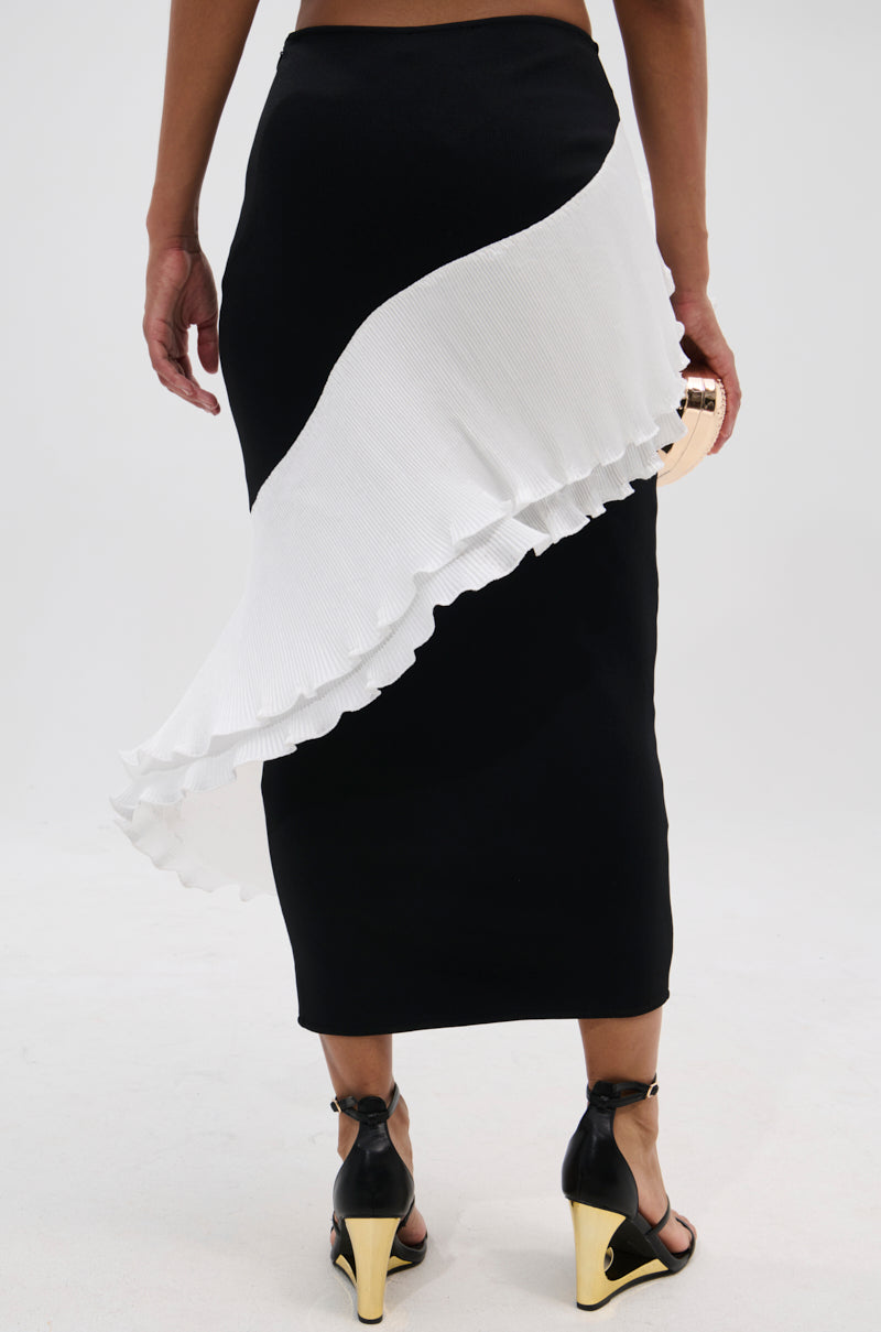 A person wearing the FASHION ICON RUFFLE BANDAGE MAXI SKIRT, featuring black pleats and a white ruffled diagonal panel, paired with black high-heeled sandals with gold accents, stands against a plain light background. 5