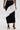 A person wearing the FASHION ICON RUFFLE BANDAGE MAXI SKIRT, featuring black pleats and a white ruffled diagonal panel, paired with black high-heeled sandals with gold accents, stands against a plain light background.