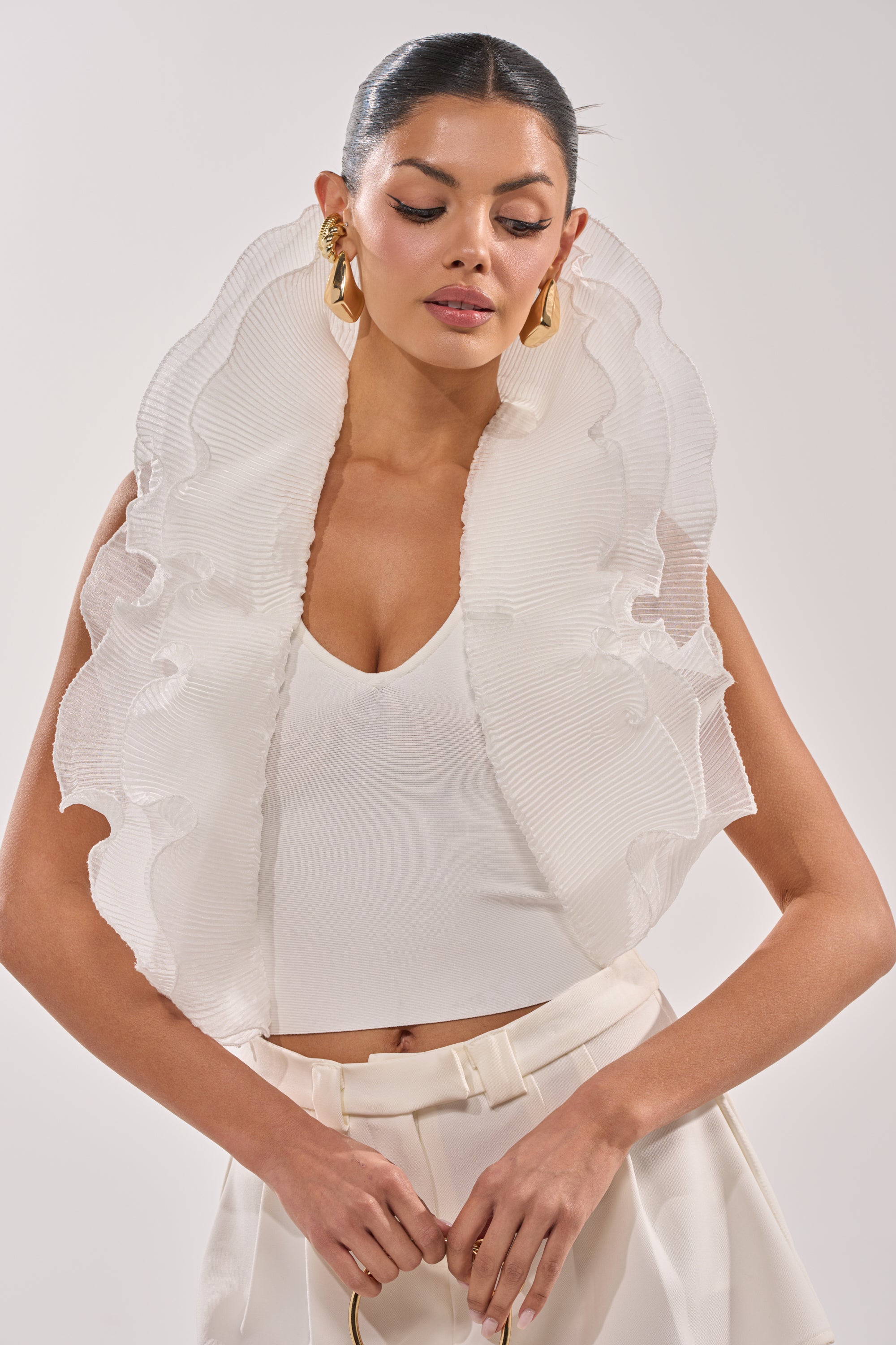 A woman wears the EXTRA BANDAGE RUFFLE BLOUSE featuring a sculptural neckline and ruffle details, paired with high-waisted trousers and gold earrings, standing against a plain background. 1