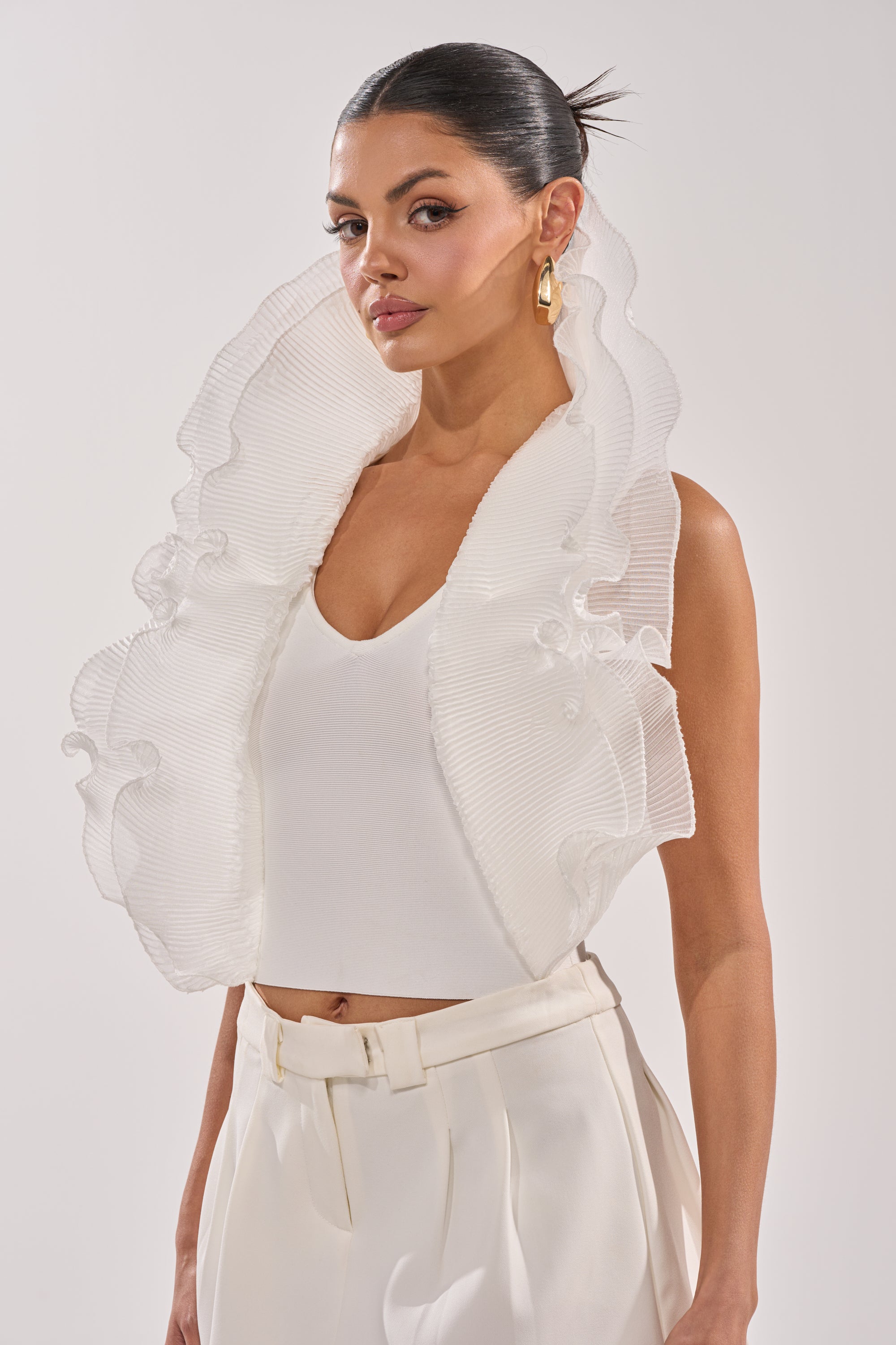 A woman in the EXTRA BANDAGE RUFFLE BLOUSE with a sculptural neckline, white high-waisted pants, gold hoop earrings, and a translucent ruffled collar poses against a plain background. 0