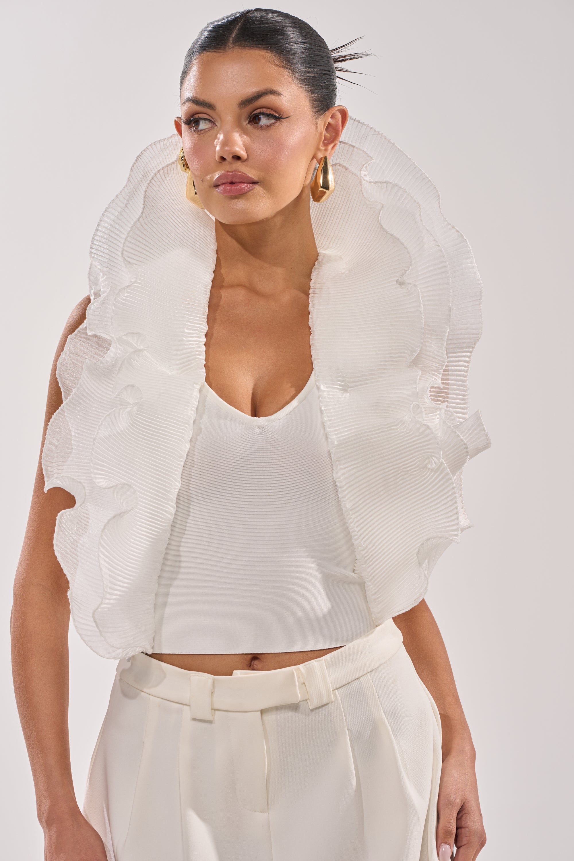 A woman with slicked-back hair wears gold hoop earrings, high-waisted white pants, and the EXTRA BANDAGE RUFFLE BLOUSE—a sleeveless white top with dramatic semi-transparent pleated ruffles framing her shoulders. 3