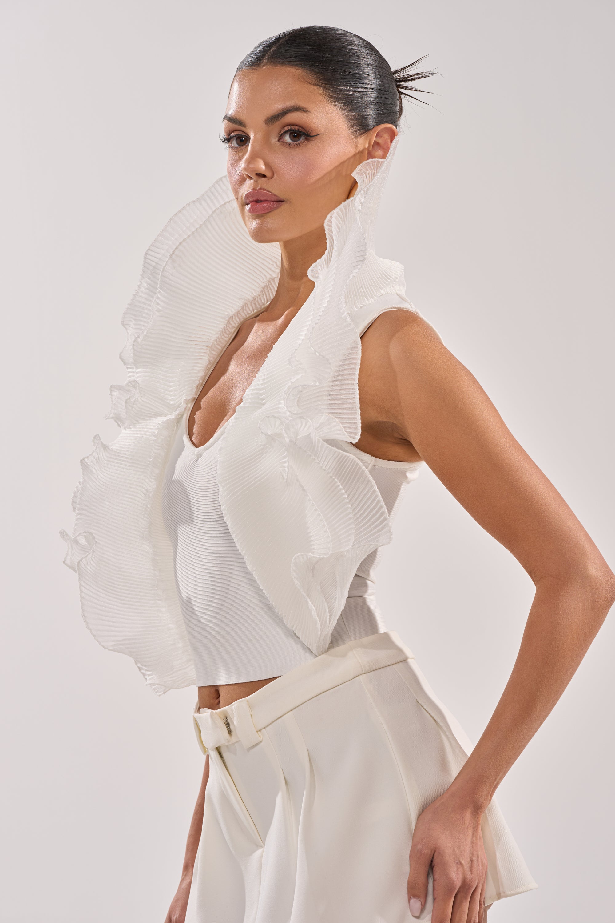 A woman stands confidently against a light background, wearing the EXTRA BANDAGE RUFFLE BLOUSE—white with dramatic translucent organza ruffles—and high-waisted white pants, her hair slicked back as she looks at the camera. 4