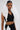 A person wears the FASHION ICON RUFFLE BANDAGE BLOUSE, a black sleeveless crop top with a dramatic, 3D white pleated ruffle collar extending from the shoulders, paired with a high-waisted skirt against a plain light background.