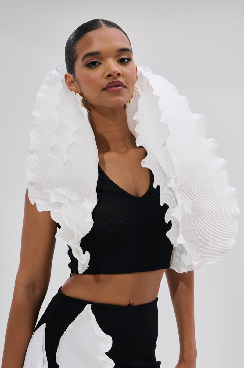 A woman in slicked-back hair and gold earrings wears the FASHION ICON RUFFLE BANDAGE BLOUSE— a black crop top and skirt set with bold, sculptural 3D ruffle collar accents at her shoulders and waist, standing against a light background. 1