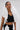 A woman in slicked-back hair and gold earrings wears the FASHION ICON RUFFLE BANDAGE BLOUSE— a black crop top and skirt set with bold, sculptural 3D ruffle collar accents at her shoulders and waist, standing against a light background.