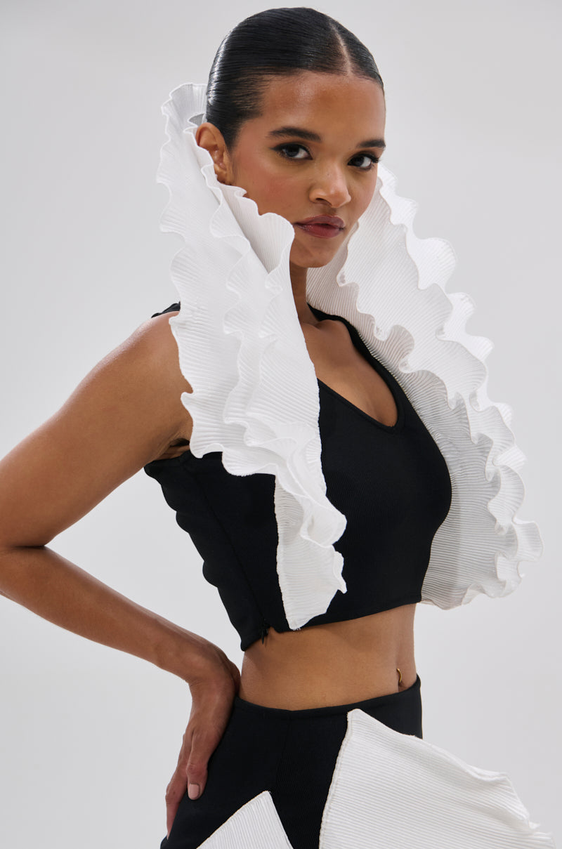 A woman with slicked-back hair wears the FASHION ICON RUFFLE BANDAGE BLOUSE, featuring sculptural 3D ruffle collar details at the shoulders, styled against a plain light background. 2