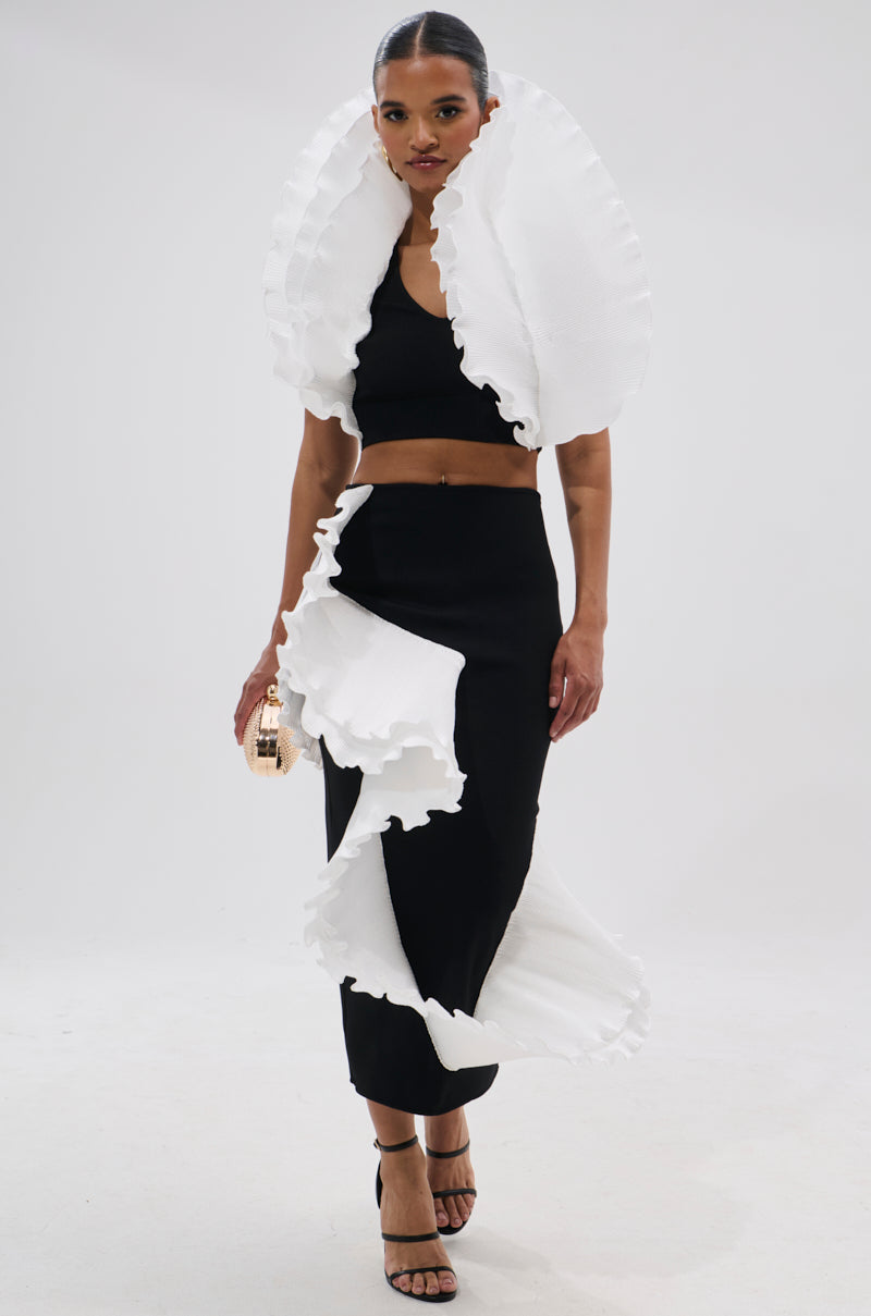 A woman models the FASHION ICON RUFFLE BANDAGE BLOUSE, a bold black crop top featuring large white 3D ruffle collars. She pairs it with a matching skirt, gold clutch, and black strappy heels against a plain white backdrop. 5