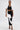 A woman wears the FASHION ICON RUFFLE BANDAGE BLOUSE, a striking black stretch-knit top with dramatic white ruffled accents on the shoulders, paired with a matching skirt and black strappy heels, standing against a plain white background.
