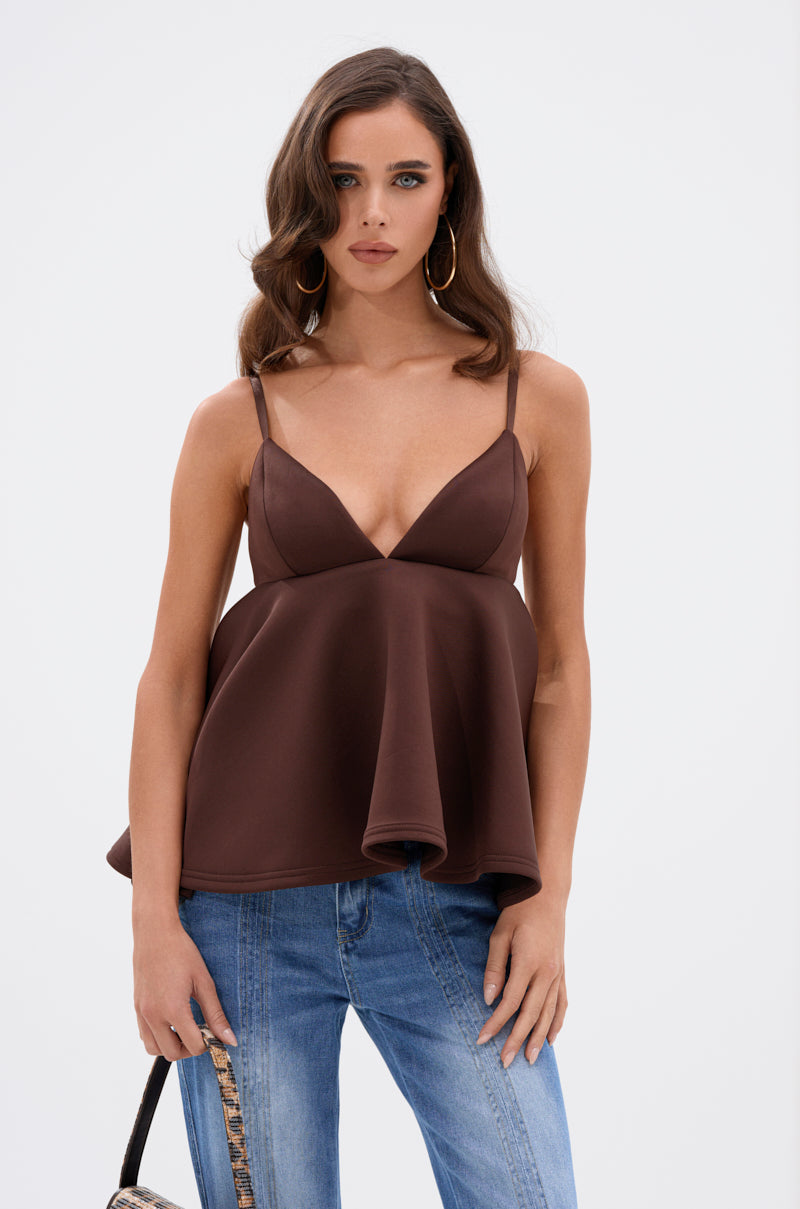 A woman with wavy brown hair wears the BIBI SCUBA CAMI TOP IN BROWN, a sleeveless deep V-neck babydoll-style top, paired with blue jeans. She stands against a white background, holding a patterned bag and wearing large hoop earrings. 0