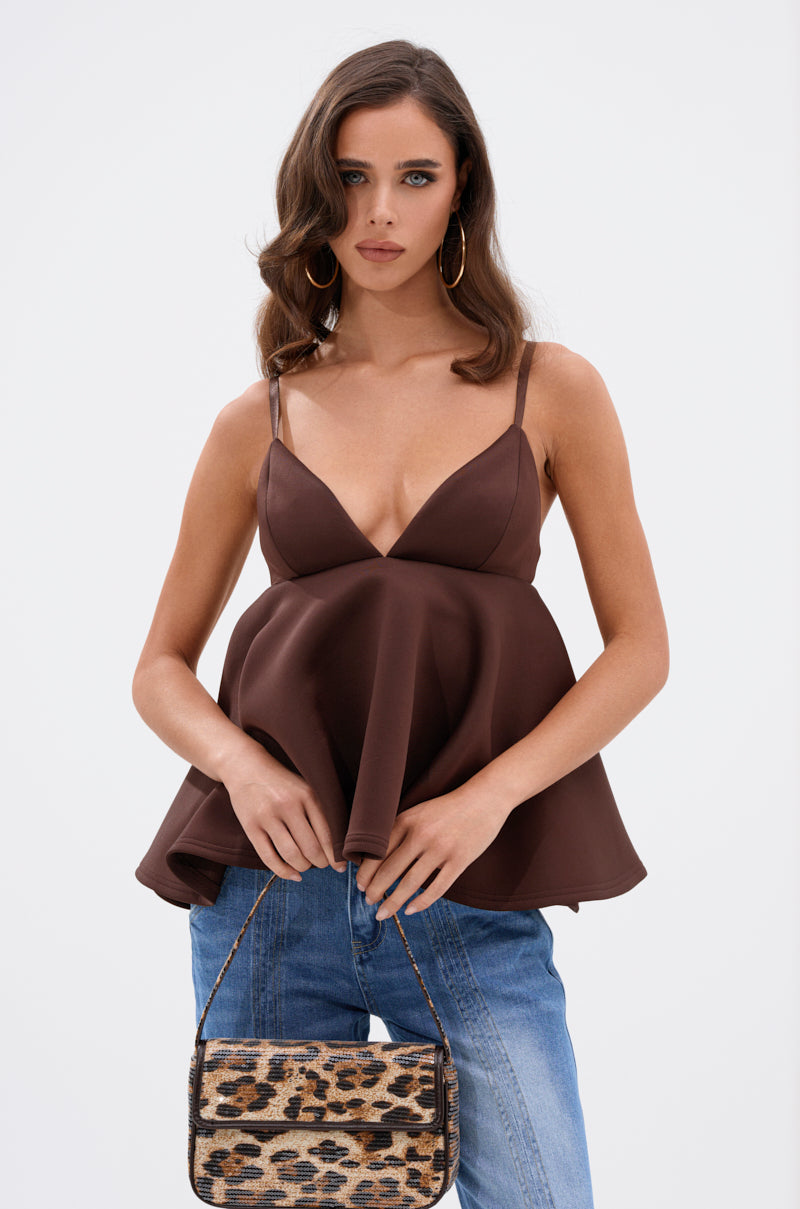 A woman with wavy brown hair wears the BIBI SCUBA CAMI TOP IN BROWN with a babydoll silhouette, blue jeans, hoop earrings, and holds a small leopard-print handbag against a plain white background. 1