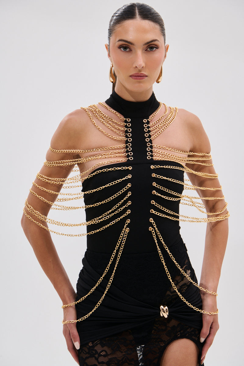 A woman with slicked-back hair wears the CHAINS DRIP BODYSUIT, a black piece with a bold silhouette and gold-tone chain details draped over her shoulders, chest, and arms. She stands against a plain light background, gazing directly at the camera. 0