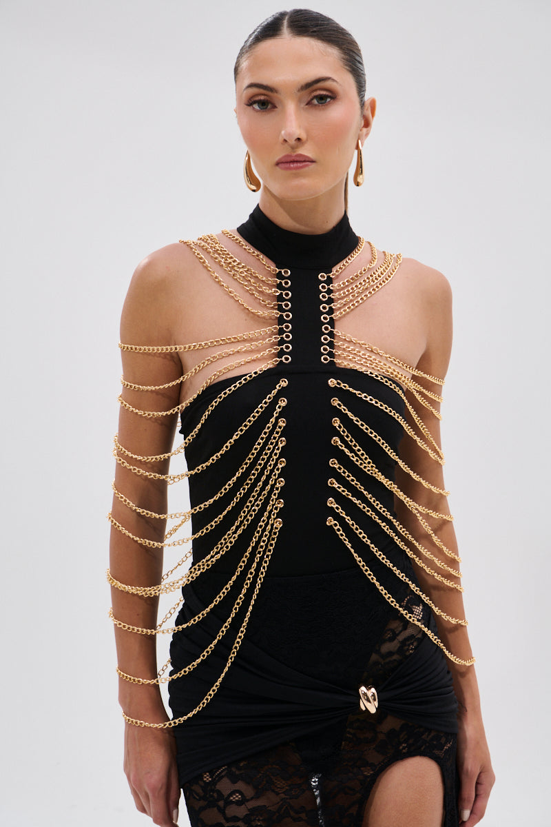 A woman with slicked-back hair and hoop earrings exudes confidence in the CHAINS DRIP BODYSUIT—a sleeveless black piece accented by gold-tone chains draped over her shoulders—posed against a plain light background. 1