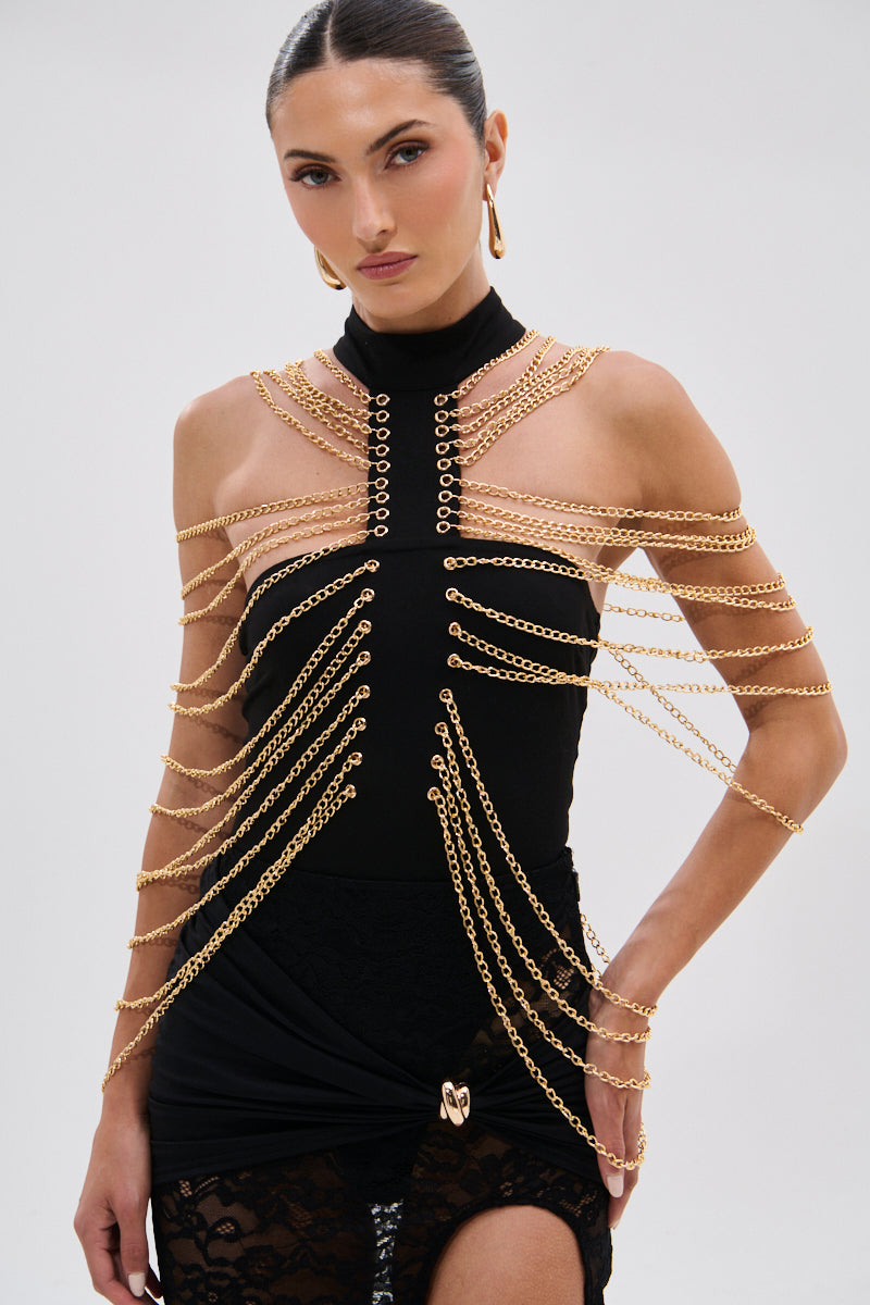 A woman with dark hair pulled back models the CHAINS DRIP BODYSUIT, featuring gold-tone chain details draped over her shoulders, chest, and arms. She pairs it with gold hoop earrings against a plain light background. 2