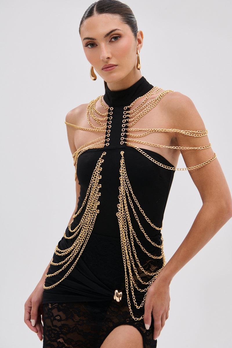 In the CHAINS DRIP BODYSUIT, a woman stuns in a black high-neck, sleeveless piece with gold-tone chain details at the shoulders and hips. With slicked-back hair and gold hoop earrings, she poses confidently with one hand on her leg. 3