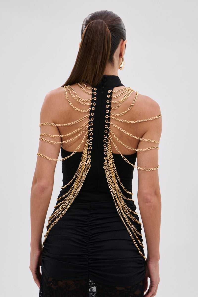 A woman with long, straight hair wears the CHAINS DRIP BODYSUIT, a black piece with high-impact silhouette, gold-tone chains draped across her back and shoulders, a high neckline, and ruched detailing for an elegant finish. 6