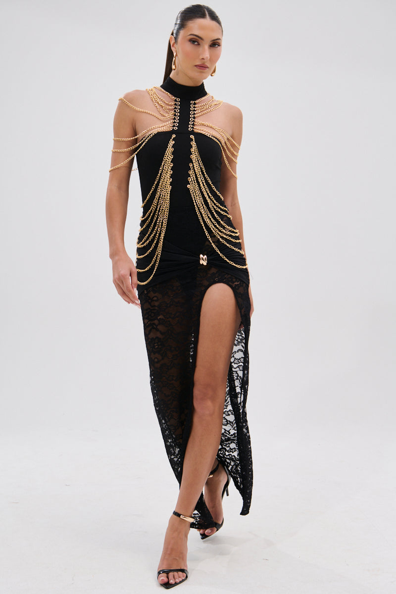 A woman wearing the CHAINS DRIP BODYSUIT, featuring black lace, a striking silhouette, and gold-tone chain accents, strides confidently in high-heeled sandals against a plain white background. 7