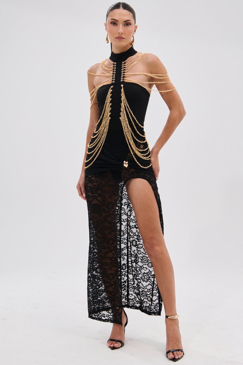 A woman wears the CHAINS DRIP BODYSUIT, a fitted black piece with a lace skirt, high slit, and gold-tone chain details over the shoulders and bodice, styled with black high-heeled sandals against a plain light background. 8