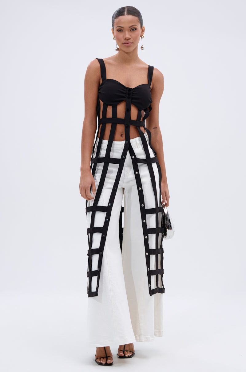 A woman with slicked-back hair and hoop earrings wears the JULIETTE BRALETTE CAGE TOP, featuring padded cups and a cage overlay, styled with white wide-leg pants, black heels, and a small handbag against a plain background.