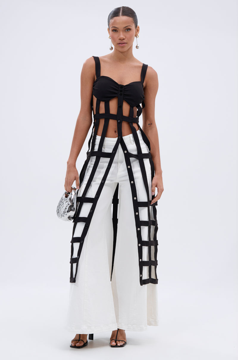 A woman faces forward wearing the JULIETTE BRALETTE CAGE TOP in black over a black crop top and wide-leg white pants. She holds a small patterned handbag, wears black sandals, and has her hair pulled back.
