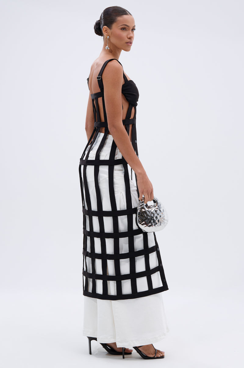 A woman wearing the JULIETTE BRALETTE CAGE TOP over a white floor-length skirt, holding a small silver bag, with her hair in a neat bun and statement earrings, stands in profile against a plain background.