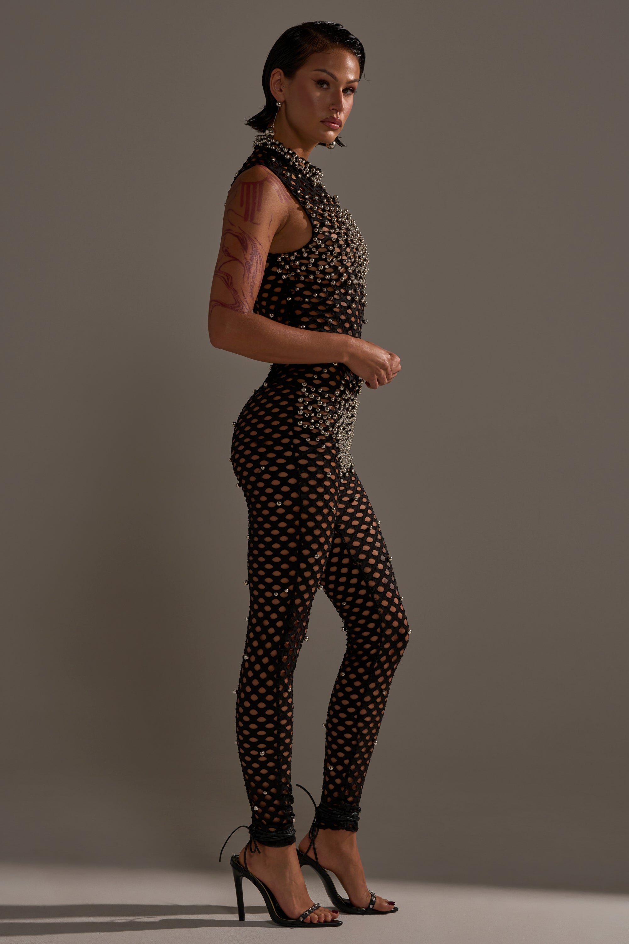 A person stands in profile against a gray background, wearing the COULD NEVER BE ME BEADED MESH JUMPSUIT featuring a textured circular pattern, paired with black high-heeled sandals and showing visible arm tattoos.