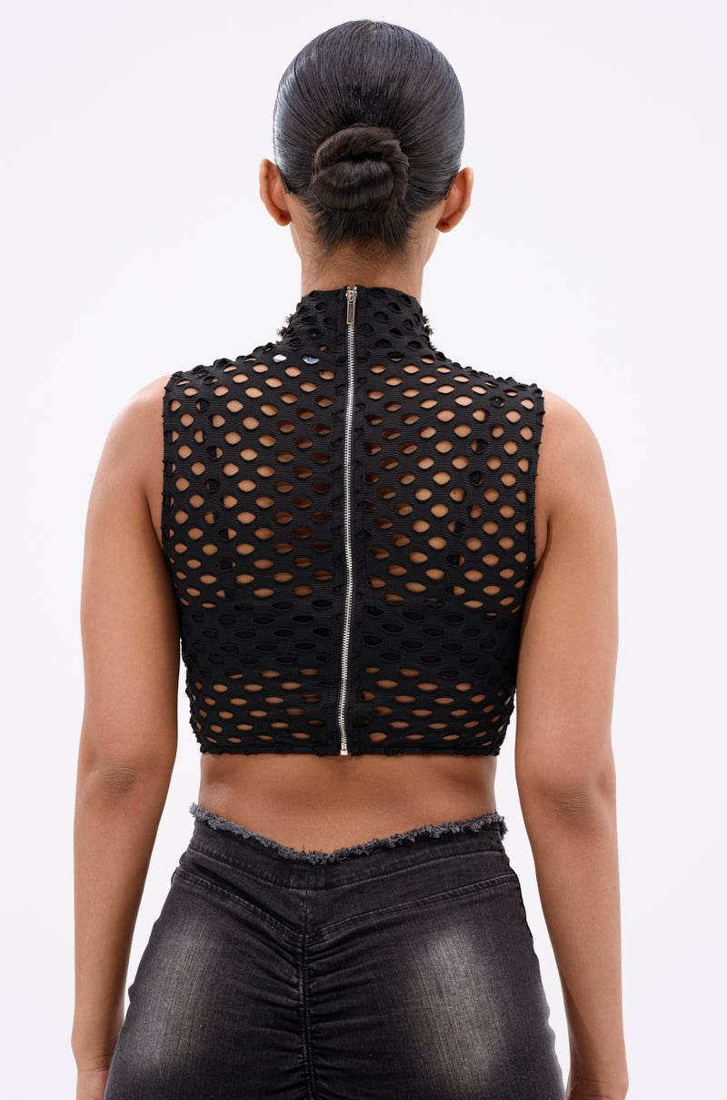 A woman with her hair in a neat bun faces away, wearing the METAL ORB DISTRESSED SLEEVELESS TOP—black, high-neck, accented with silver-tone studs and a back zipper—paired with dark high-waisted jeans.