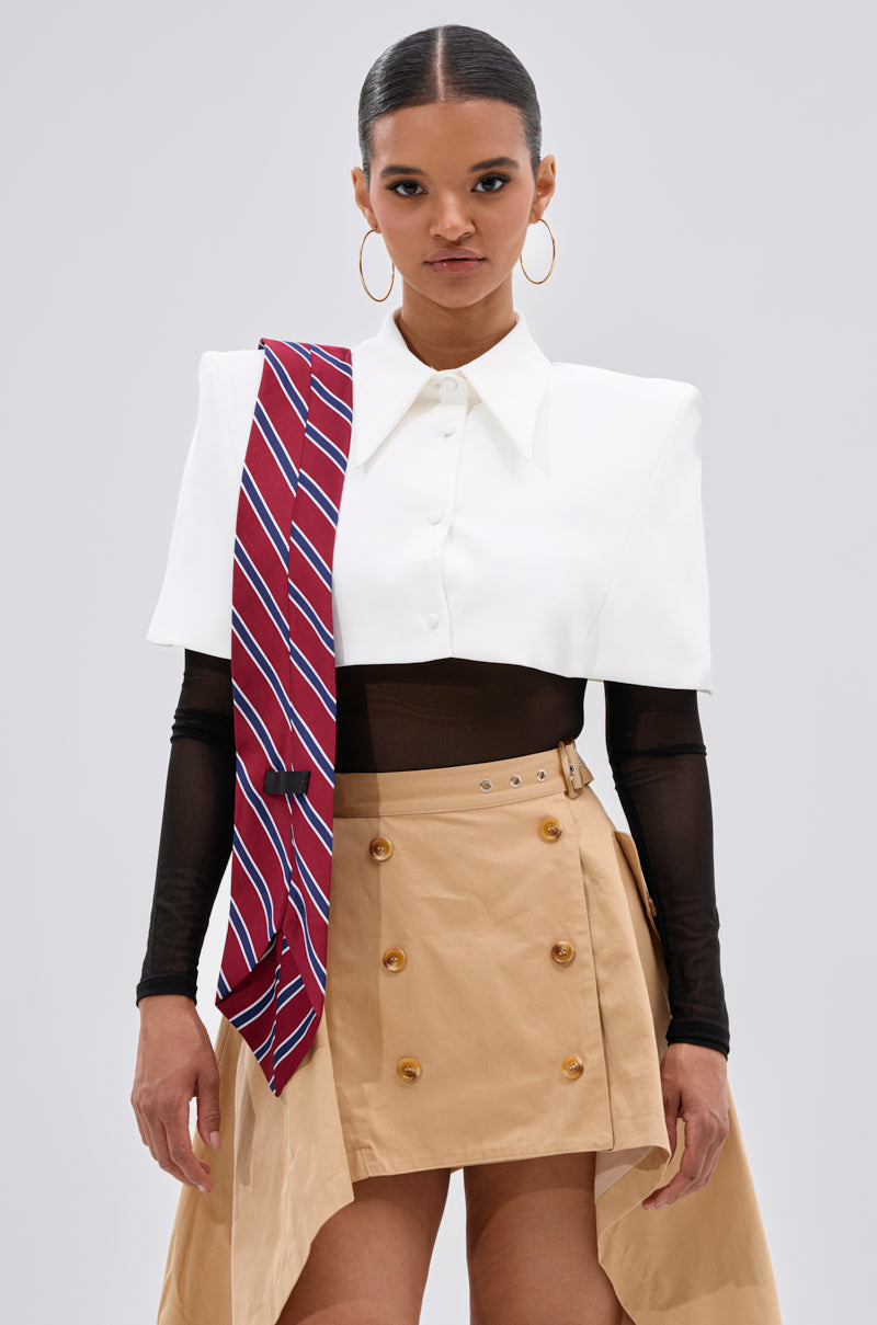 A woman faces forward in the GET WITH IT BUTTON DOWN BODYSUIT, styled with a white capelet, tan asymmetrical skirt with buttons and belt, red-striped tie over one shoulder, hoop earrings, and her hair pulled back.