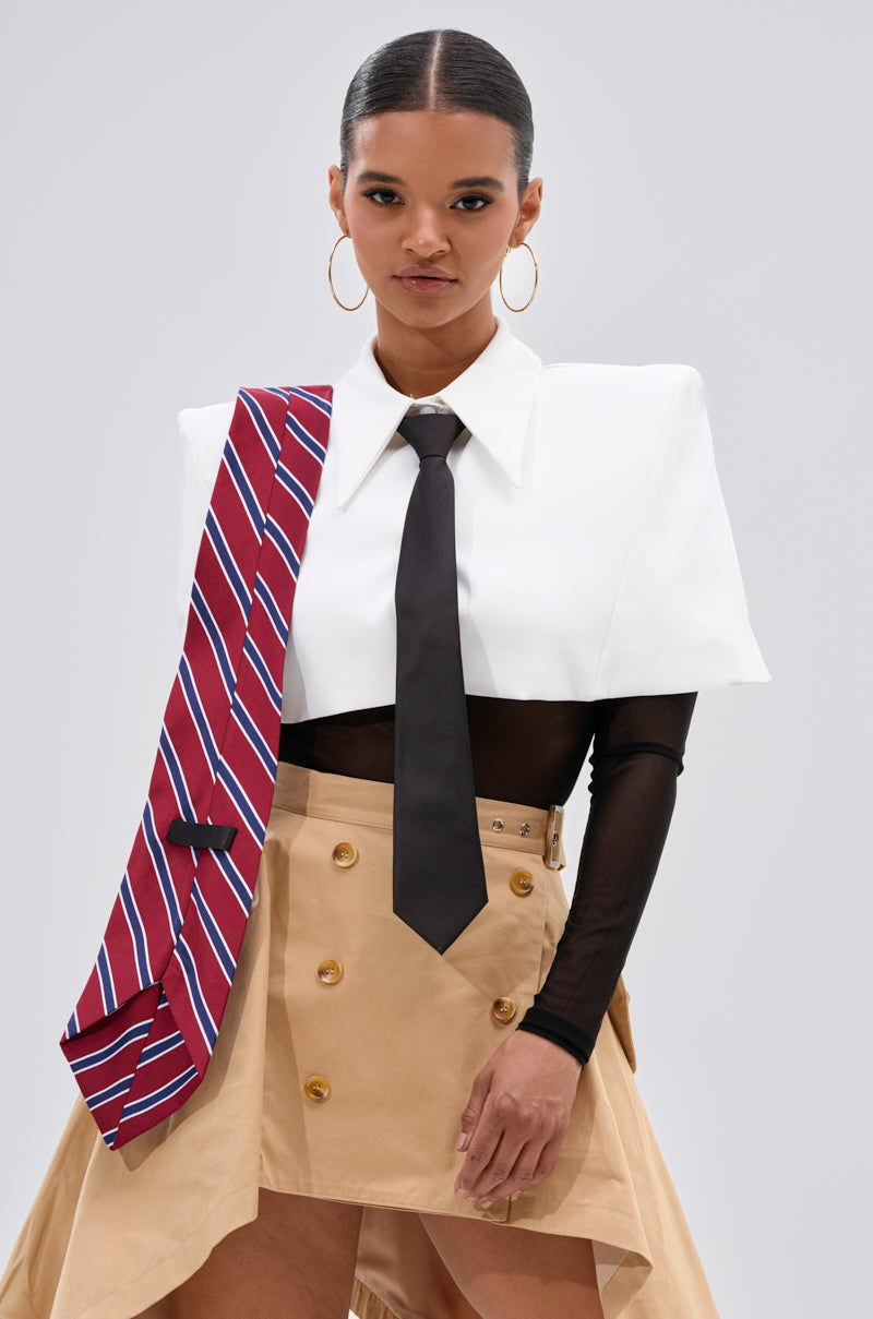 A woman poses confidently in the white GET WITH IT BUTTON DOWN BODYSUIT styled over a black mesh shirt, black tie, beige asymmetrical skirt with gold buttons, and a red striped tie. She completes the look with hoop earrings and sleek hair.