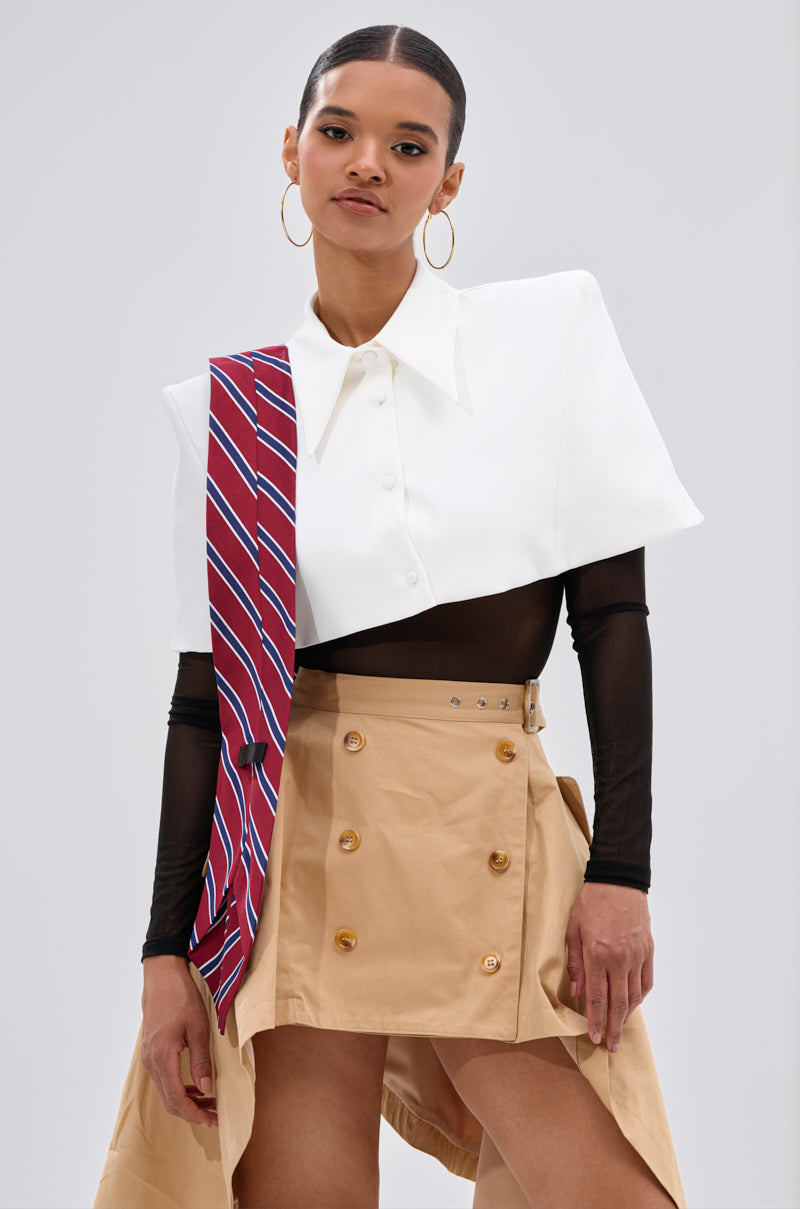 A woman stands against a plain light background wearing the GET WITH IT BUTTON DOWN BODYSUIT styled with a white cropped capelet overlay, sheer black top, tan asymmetrical skirt with buttons, and a red-striped tie draped over one shoulder.