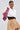 A woman in the GET WITH IT BUTTON DOWN BODYSUIT and an asymmetrical tan skirt with buttons poses with a red striped necktie over her shoulder. She accessorizes with large hoop earrings and has her hair slicked back against a plain background.