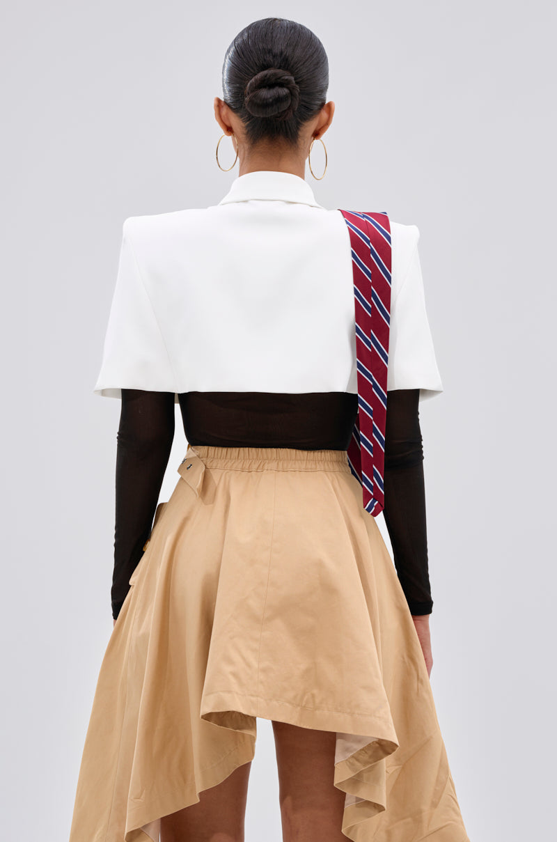 A woman faces away, wearing the GET WITH IT BUTTON DOWN BODYSUIT styled with a white cropped blazer featuring a capelet overlay, a red striped tie over one shoulder, tan high-low skirt, large hoop earrings, and her hair in a sleek bun.