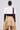 A woman faces away, wearing the GET WITH IT BUTTON DOWN BODYSUIT styled with a white cropped blazer featuring a capelet overlay, a red striped tie over one shoulder, tan high-low skirt, large hoop earrings, and her hair in a sleek bun.