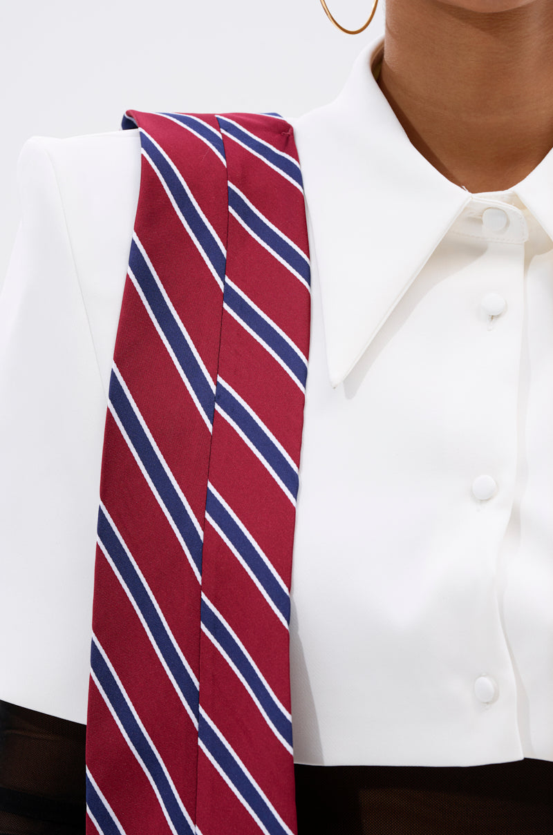 Wearing the GET WITH IT BUTTON DOWN BODYSUIT, this person pairs gold hoop earrings and a striped red, blue, and white necktie over their shoulder, highlighted by a stylish capelet overlay for an extra touch of flair.