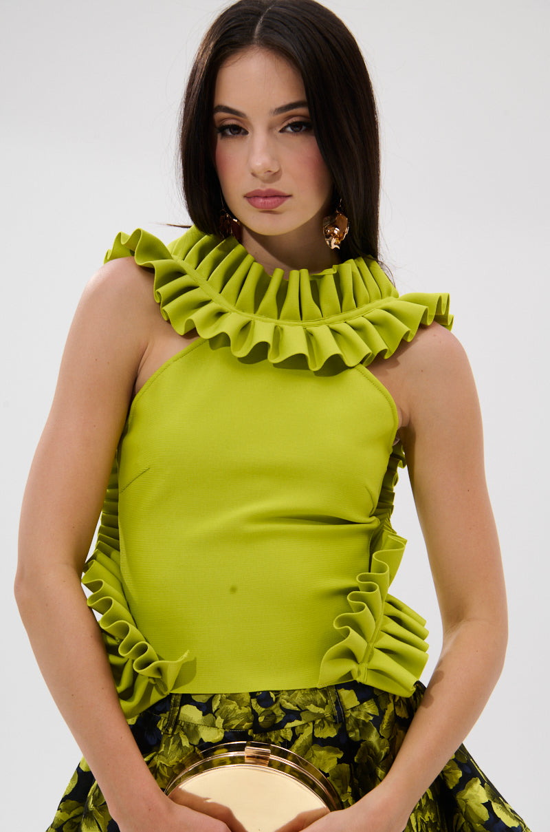 A woman with long dark hair wears the BAROQUE MOCK NECK TOP IN CHARTREUSE, featuring dramatic pleated ruffles, paired with a floral skirt and holding a round gold purse, gazing at the camera against a plain background.
