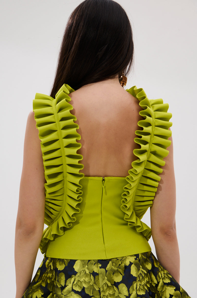 A woman with long dark hair faces away, wearing the Baroque Mock Neck Top in chartreuse with dramatic ruffled trim on the shoulders and back, paired with a skirt featuring a green floral pattern.