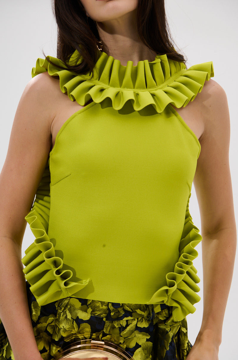 A woman wears the BAROQUE MOCK NECK TOP IN CHARTREUSE, a sleeveless top with large ruffles around the neck, armholes, and sides. Only her upper body is shown.