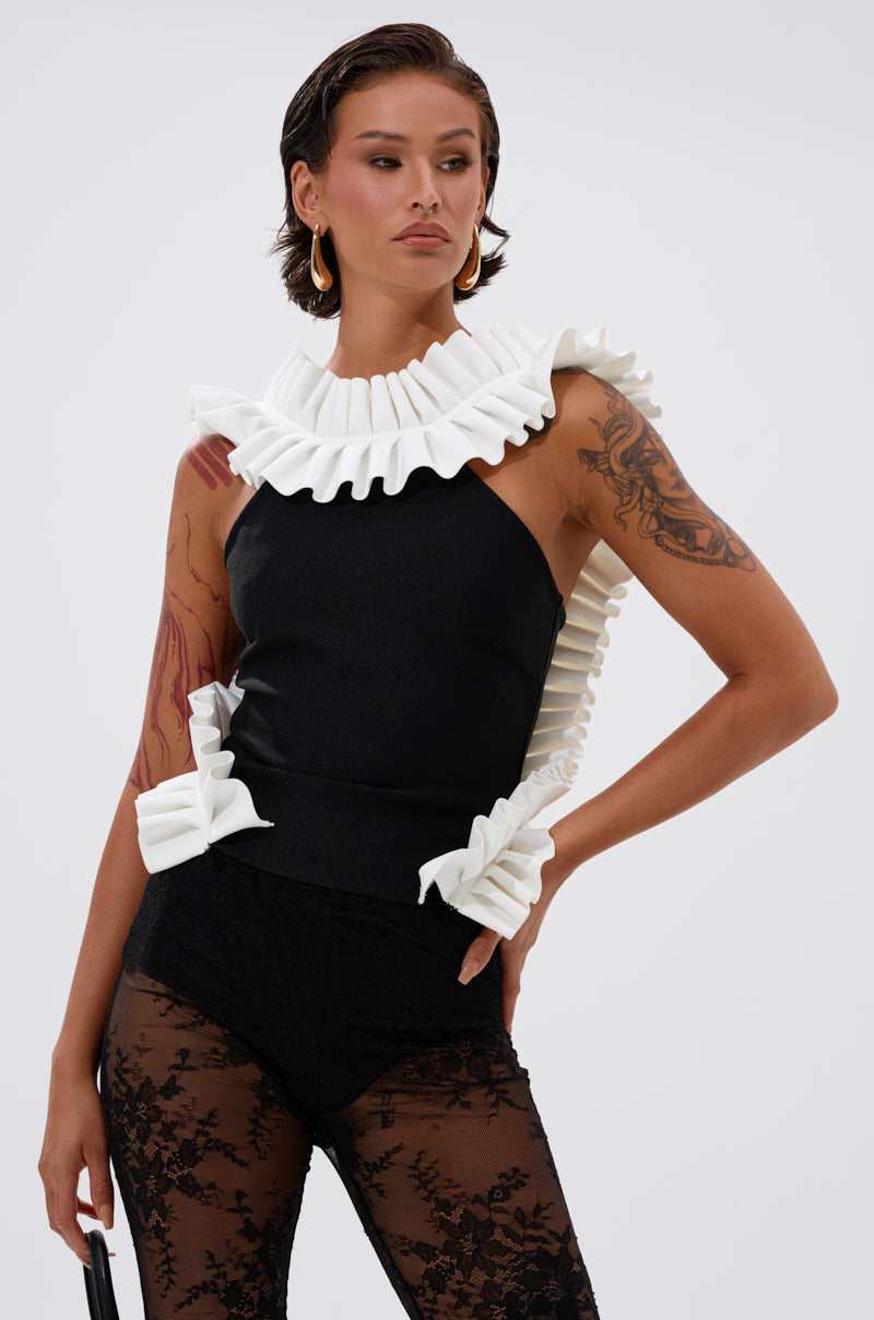 A woman models the BAROQUE MOCK NECK TOP with bold white ruffled trim at the neck, arms, and waist, paired with sheer black lace pants. She has short hair, gold hoop earrings, and visible tattoos on her left arm. 0