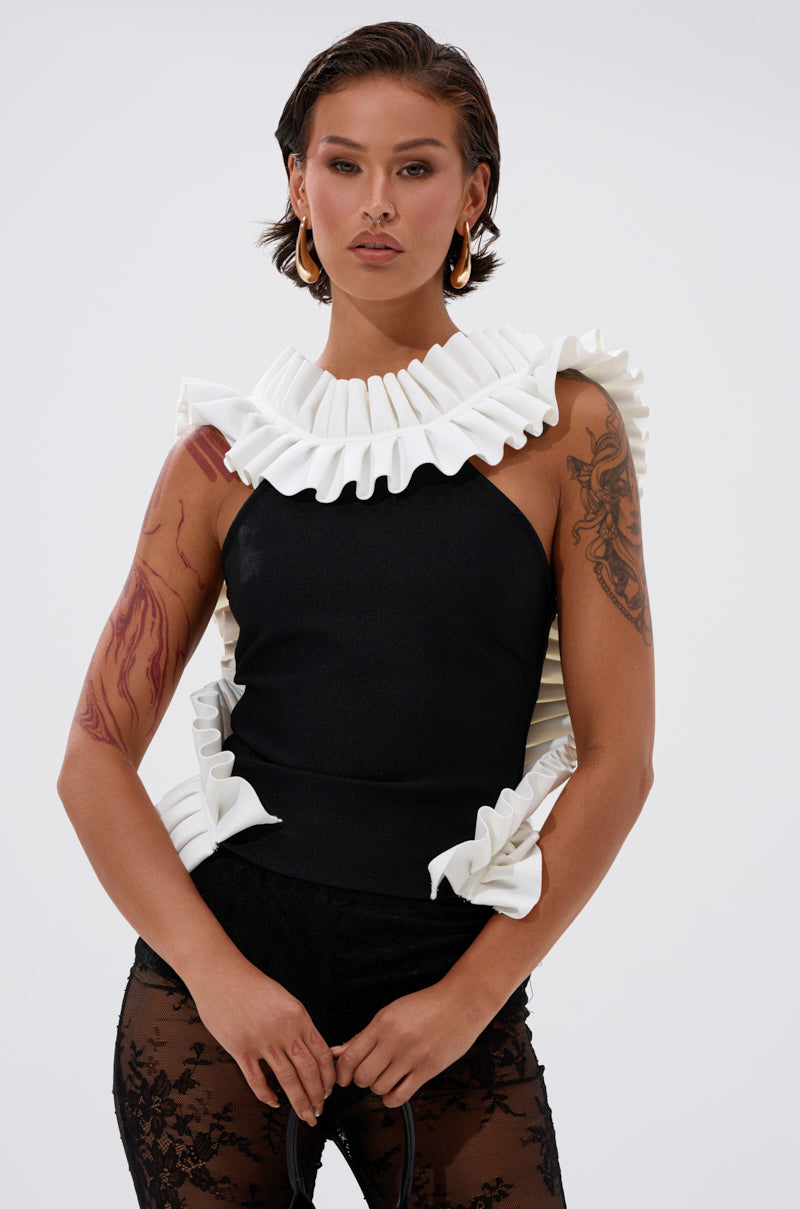 A woman with wet-look hair wears the BAROQUE MOCK NECK TOP, featuring dramatic white ruffles at the collar and sides, paired with sheer black lace pants. She stands confidently against a plain background. 1