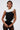 A woman with wet-look hair wears the BAROQUE MOCK NECK TOP, featuring dramatic white ruffles at the collar and sides, paired with sheer black lace pants. She stands confidently against a plain background.