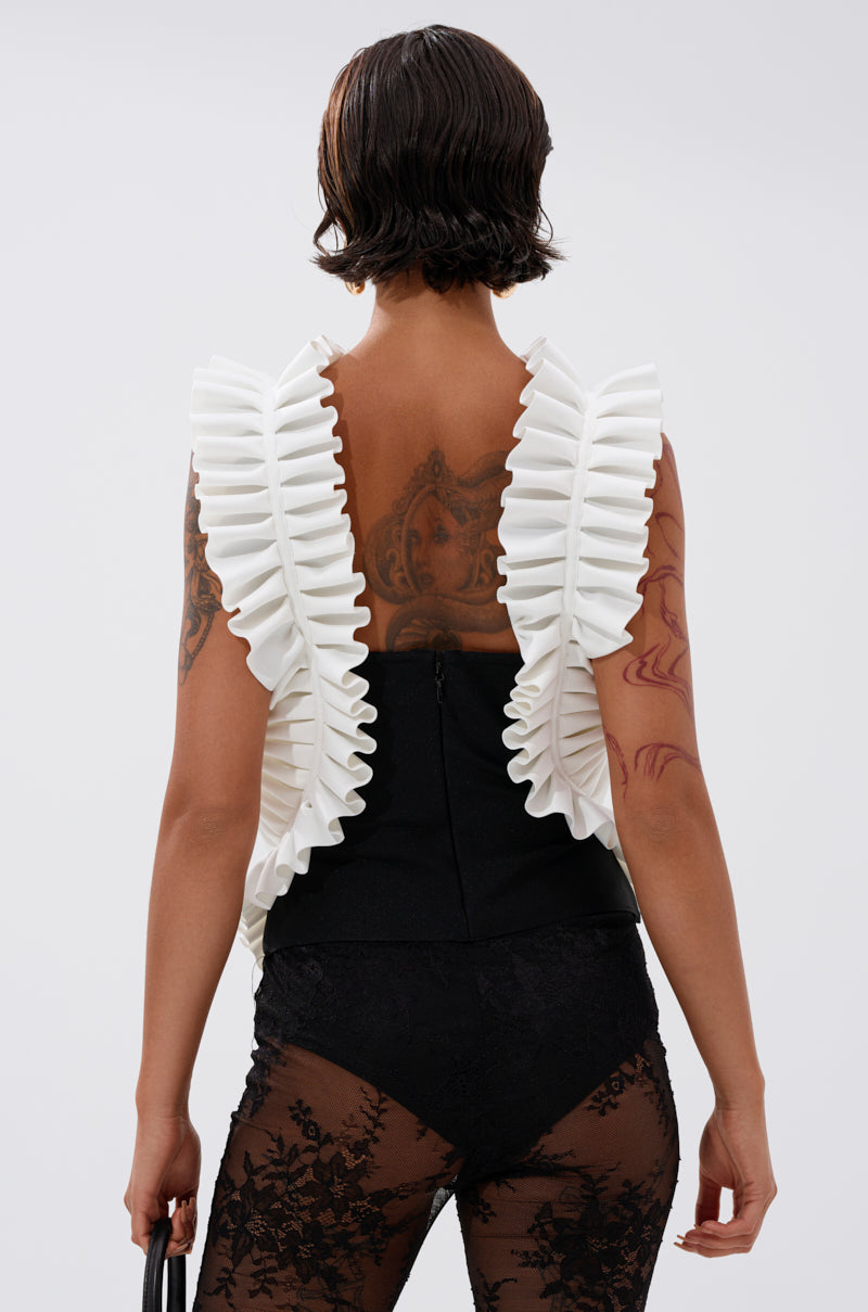 A person with short dark hair and tattoos is seen from behind, wearing the BAROQUE MOCK NECK TOP—a vintage-inspired sleeveless white ruffled piece—over a black outfit with sheer lace detailing. They hold a small black item at their side. 3