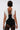 A person with short dark hair and tattoos is seen from behind, wearing the BAROQUE MOCK NECK TOP—a vintage-inspired sleeveless white ruffled piece—over a black outfit with sheer lace detailing. They hold a small black item at their side.