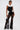 A person poses confidently in the BAROQUE MOCK NECK TOP featuring dramatic white ruffles, paired with sheer black lace flared pants, gold pointed shoes, and a small black handbag against a plain white background.