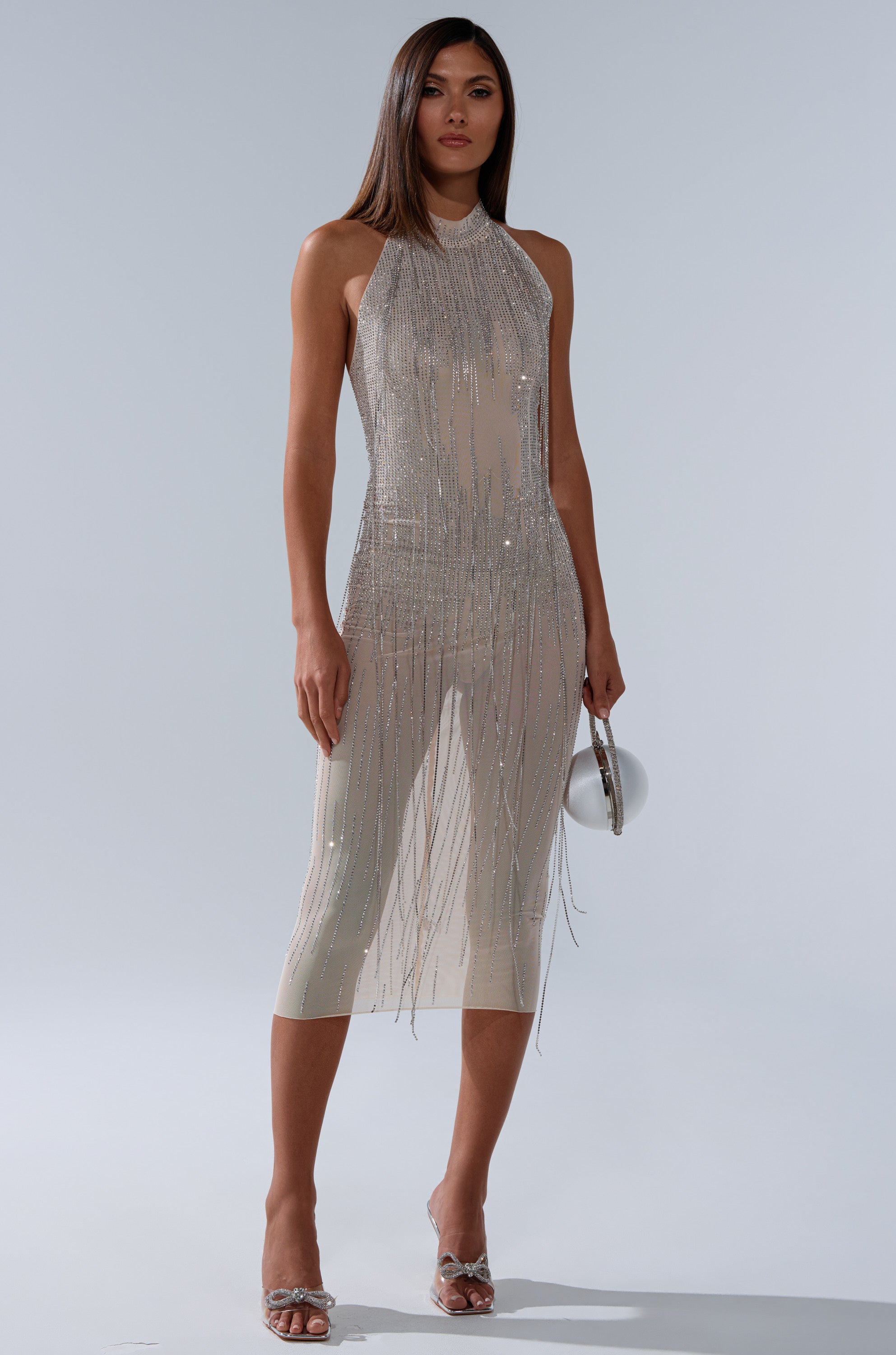 A woman stands against a light gray background wearing the sleeveless KEKE MIDI DRESS WITH RHINESTONE FRINGE, featuring silver bead and fringe embellishments. She holds a small silver bag and wears silver open-toe heels.