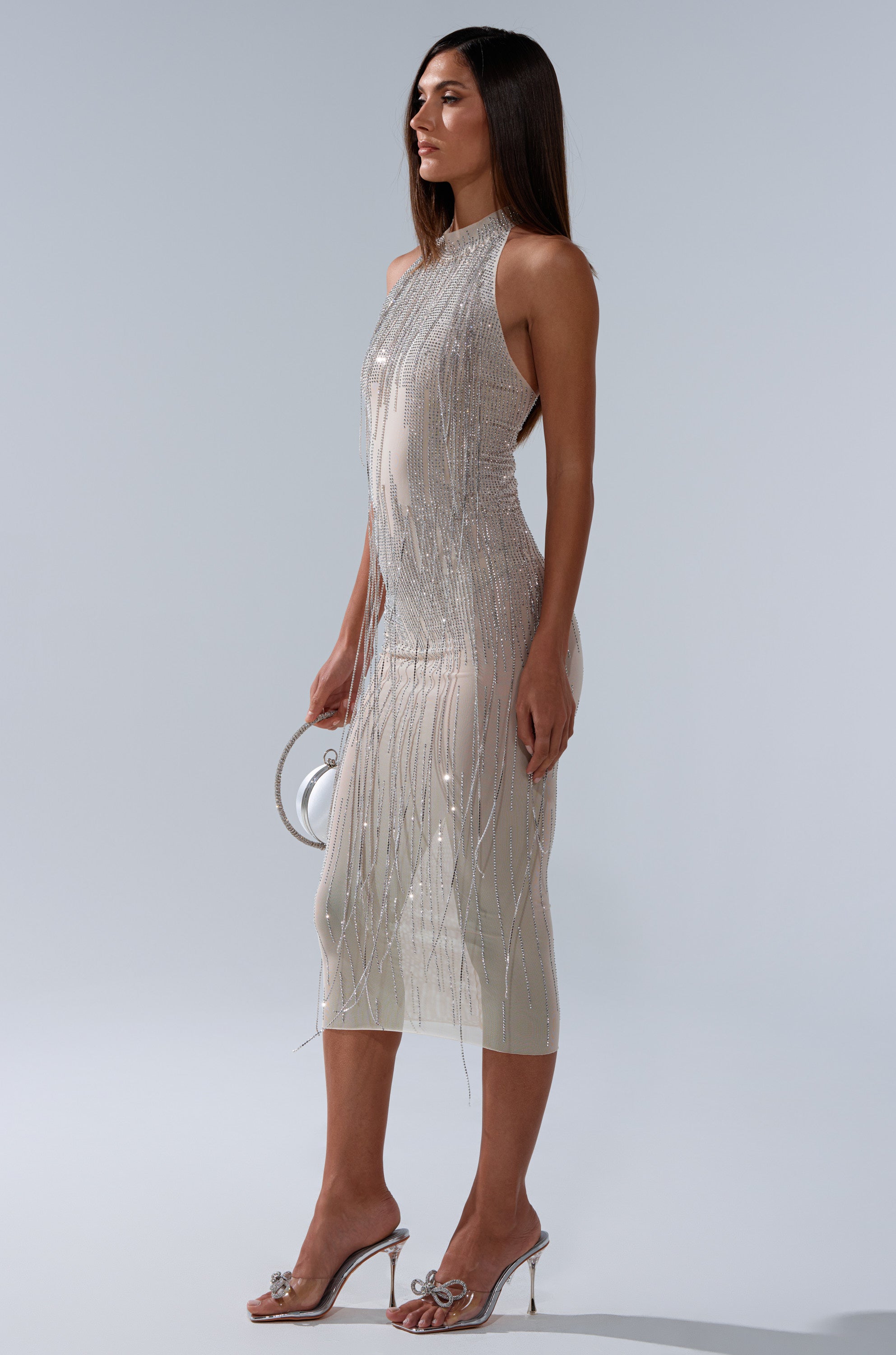 A woman in profile wears the sleeveless, fitted KEKE MIDI DRESS WITH RHINESTONE FRINGE. She holds a small round purse, pairs it with clear high heels, and stands against a plain light gray background.