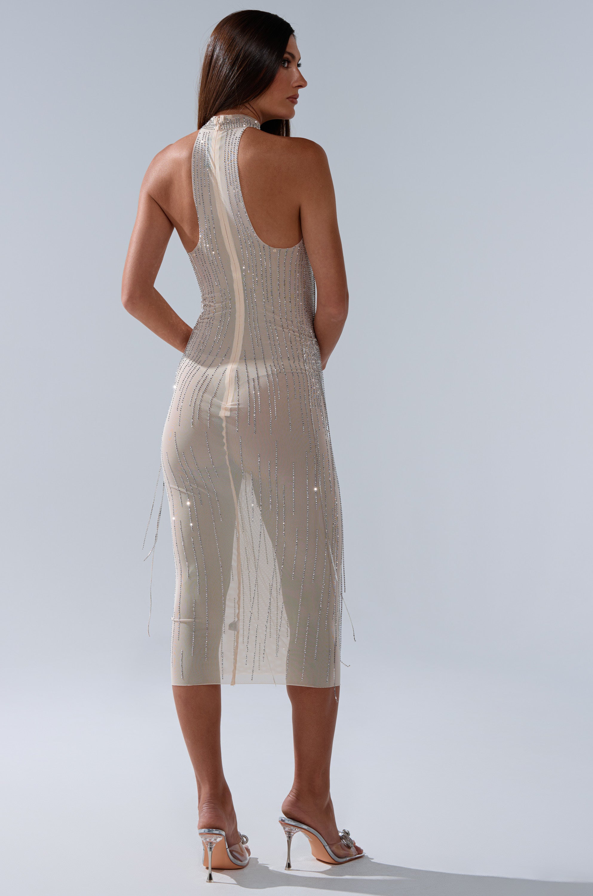 A woman with long brown hair and high-heeled sandals faces away from the camera, wearing the KEKE MIDI DRESS WITH RHINESTONE FRINGE on a neutral background.