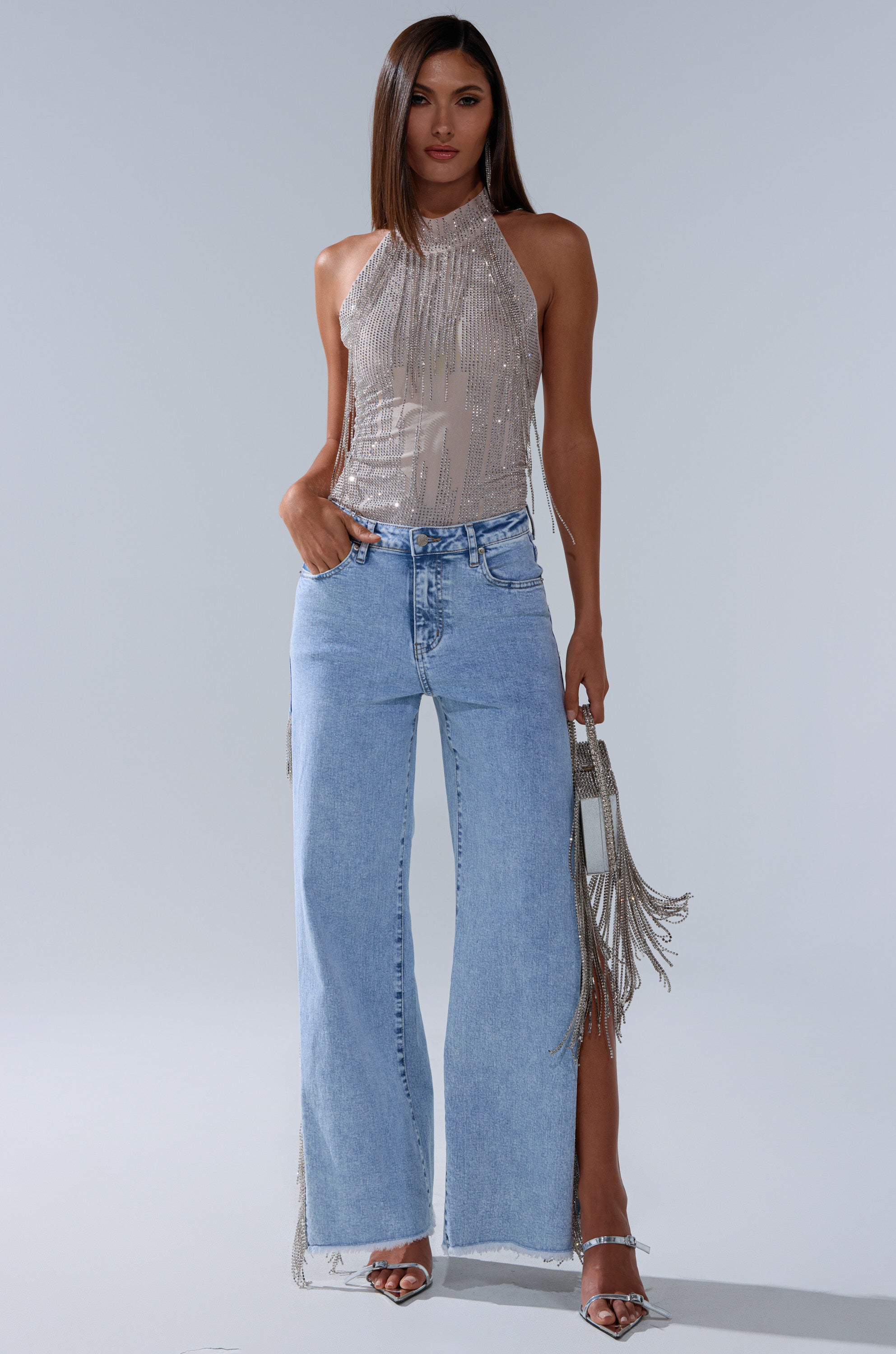 A woman stands confidently in the ALL SEASON RHINESTONE MOCK NECK TOP, paired with light blue wide-leg jeans, silver high-heeled sandals, and a silver fringe handbag against a plain light background.