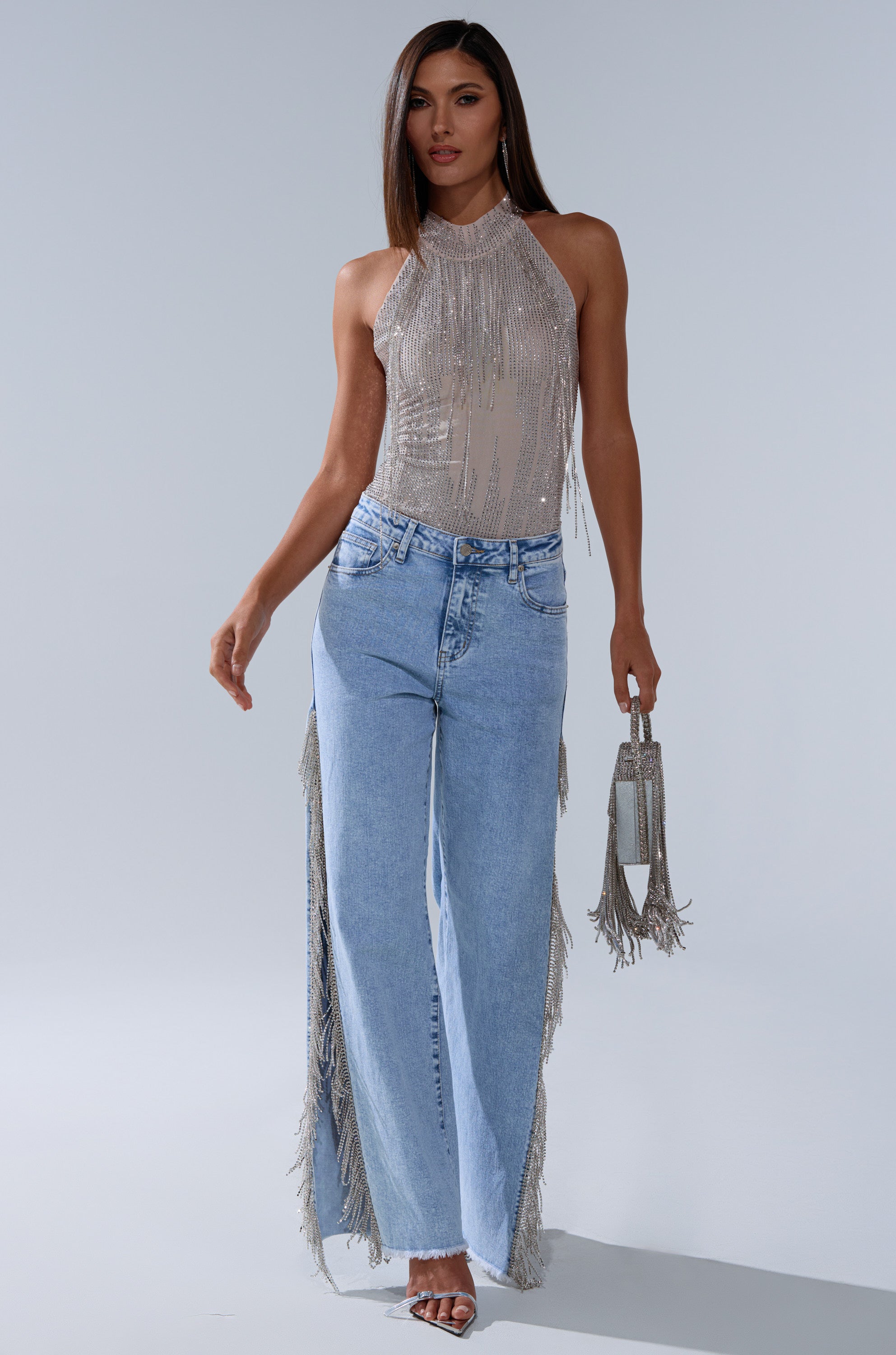 A woman poses against a plain background wearing the ALL SEASON RHINESTONE MOCK NECK TOP, paired with light blue wide-leg fringe jeans, a small silver fringe handbag, and heeled sandals.